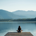 Woman standing on a wooden pier overlooking a serene lake with mountains in the background – symbolizing self-care, reflection, and expat life balance.