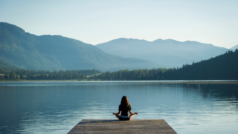 Woman standing on a wooden pier overlooking a serene lake with mountains in the background – symbolizing self-care, reflection, and expat life balance.